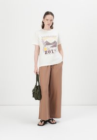 Kremfarget t-skjorte med grafisk trykk, kombinert med løse brune bukser. Outfiten er komplementert med en grønn stoffveske og svarte sandaler.