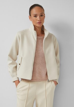 Woman wearing cream zip-up jacket over light pink knit sweater with matching cream pants, hands in pockets, against plain gray background.