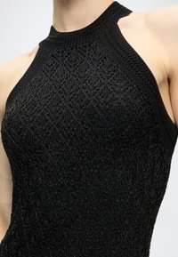 Gros plan d'une personne portant un top en maille noire sans manches avec un col montant et un motif en dentelle complexe.