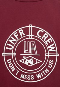 Burgundijska majica s velikim belim kružnim grafikom na kateri je napis "UNFR CREW", "UA" in obliko očesa, postavljena na gladko tkanino.