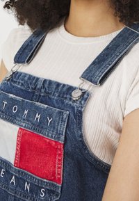 Džínové kombinézy ve tmavě modré barvě s kapsou z červeného a bílého materiálu. Obsahuje bílé vyšívané nápisy "TOMMY" a "JEANS." Viditelné stříbrné kovové detaily.