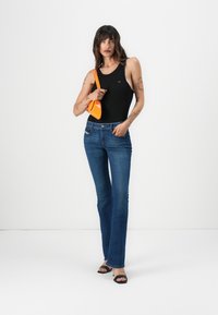 Maillot de bain noir à col haut, associé à un jean bleu évasé. Le mannequin tient un sac à main orange. Des talons ouverts noirs complètent le look.