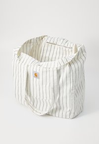 Bolsa de lona con tejido blanco y rayas verticales en azul marino. Cuenta con asas dobles y un bolsillo interior con cremallera. Parche con el logo de Carhartt visible.