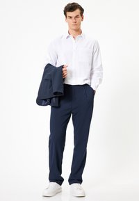 Chemise blanche à boutons avec poche, associée à un pantalon bleu marine. Le modèle tient un blazer bleu marine et porte des baskets blanches. Design simple et ajusté.