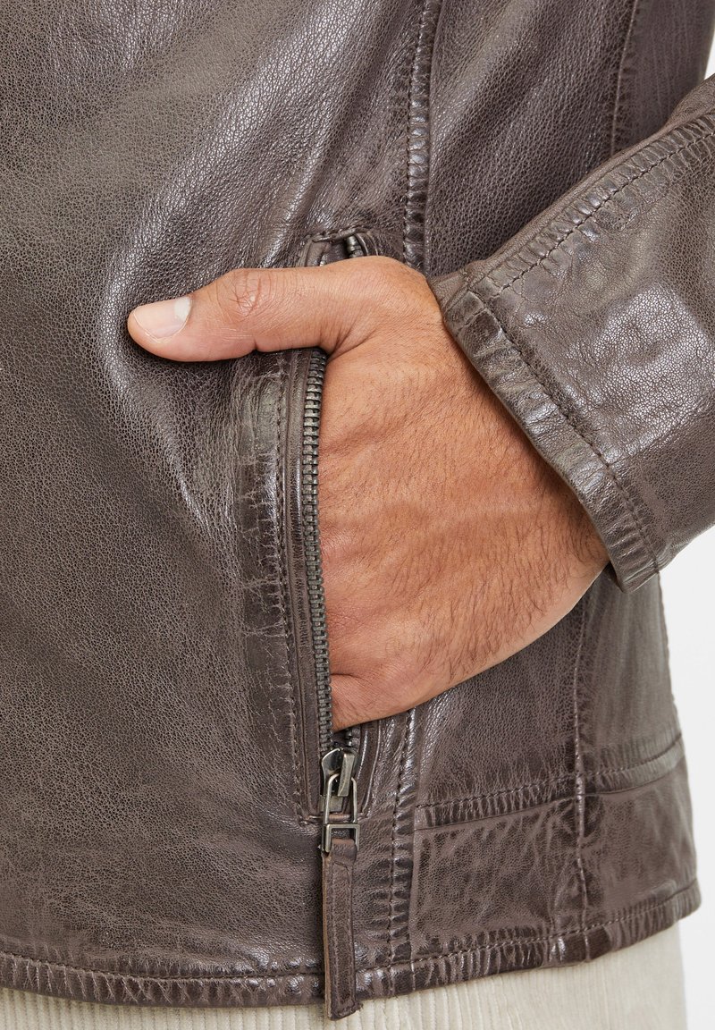 Giacca di pelle marrone con una tasca con zip e una finitura texturizzata. Una mano è poggiata nella tasca, mettendo in evidenza i dettagli di design della giacca.