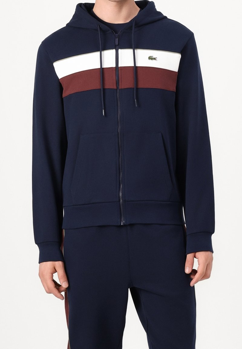Homme portant un sweat à capuche zippé bleu marine avec des bandes horizontales blanches et bordeaux sur la poitrine, assorti à un pantalon bleu marine.