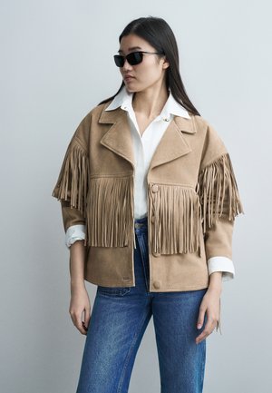 Moteris, dėvinti juodus saulės akinius, smėlio spalvos zomšos su kutais švarką ant baltos palaidinės su apykakle ir mėlynus džinsus, stovinti priešais paprastą foną.