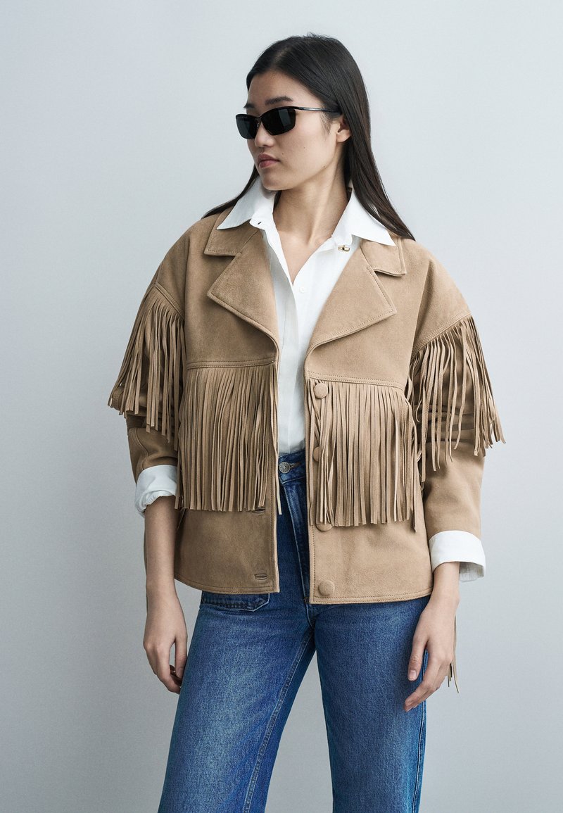 Vrouw met zwarte zonnebril, een beige suède jasje met franjes over een wit overhemd met kraag en een blauwe spijkerbroek, staand tegen een effen achtergrond.