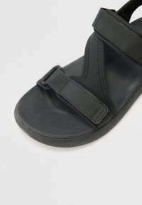 Svart sandal med tygremmar, en texturerad fotbädd och slät sula; justerbar spännare för passform.
