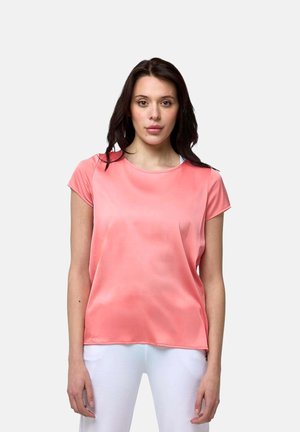 Femme aux cheveux foncés portant un haut ample corail à manches courtes et un pantalon blanc, debout devant un fond clair uni.
