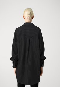 Camicia oversize a righe nere e grigie con colletto, maniche lunghe e dettaglio a pieghe sul retro. Il tessuto sembra morbido con una consistenza liscia.