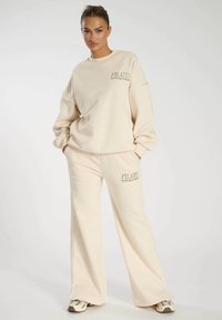 Beige Loungewear-Set bestehend aus einem lässigen Sweatshirt und weitem Hosen. Beide Teile haben eine weiche Textur und ein bedrucktes Logo-Detail.