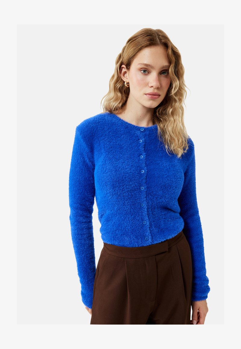 Frau mit welligem blondem Haar, die eine hellblaue, flauschige, geknöpfte Strickjacke und hoch taillierte braune Hose trägt und vor einem schlichten Hintergrund steht.