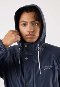 Marineblauw waterdicht jack met capuchon, drukknoopsluiting en verstelbare trekkoorden. Voorzien van een logo op de borst en elastische manchetten voor een stevige pasvorm.