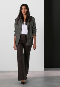 Sacou alb-negru cu model houndstooth peste un top alb, asortat cu pantaloni evazați maro și pantofi cu vârf ascuțit negri, stând pe un fundal neutru.