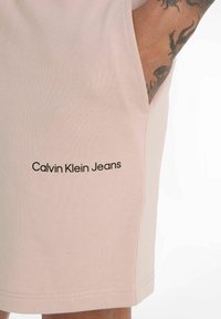 Pantalones cortos de algodón rosa con bolsillos laterales y un logo negro de "Calvin Klein Jeans" en la pierna izquierda. Textura suave y diseño casual.