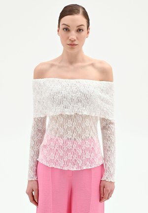 Giovane donna con i capelli tirati indietro, che indossa un top in pizzo bianco con scollo a barca e pantaloni rosa a gamba larga a vita alta, su uno sfondo neutro.