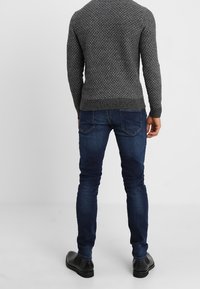 Pull en maille grise texturée, jeans slim bleu, chaussures habillées noires, vu de dos. Le pull présente des poignets et un ourlet côtelés.