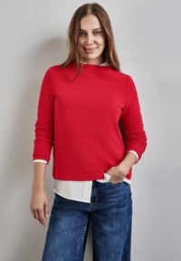 Rotes Langarmoberteil mit Rundhalsausschnitt, darunter ein weißer Kragen. Kombiniert mit blauen Denim-Jeans, glatte Textur und entspannter Schnitt.