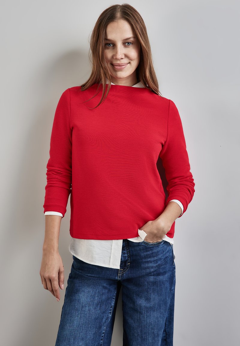 Rotes Langarmoberteil mit Rundhalsausschnitt, darunter ein weißer Kragen. Kombiniert mit blauen Denim-Jeans, glatte Textur und entspannter Schnitt.