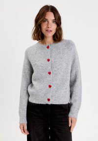 Femme aux cheveux bruns mi-longs portant un cardigan en tricot gris clair avec des boutons en forme de cœur rouges et un pantalon noir, se tenant contre un fond blanc.