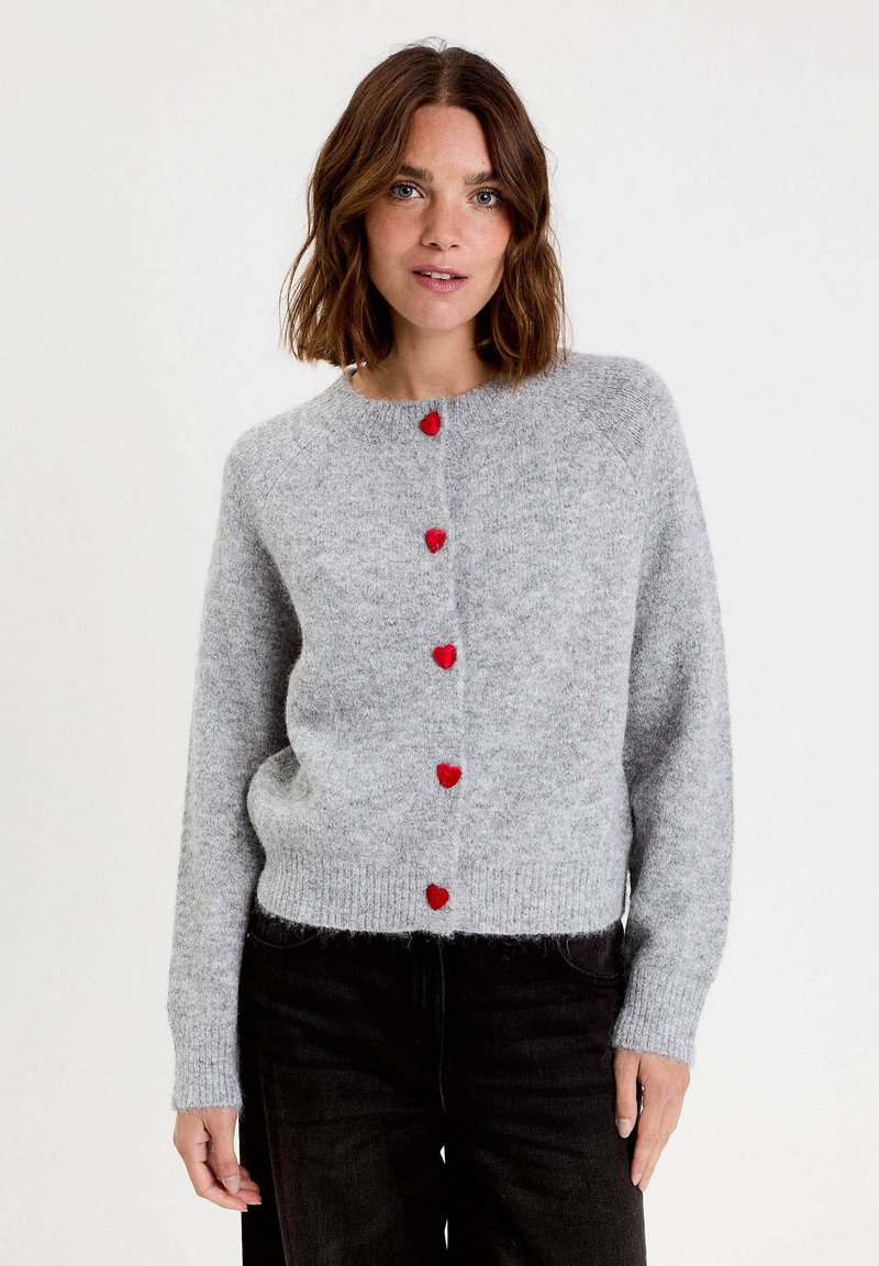 Femme aux cheveux bruns mi-longs portant un cardigan en tricot gris clair avec des boutons en forme de cœur rouges et un pantalon noir, se tenant contre un fond blanc.