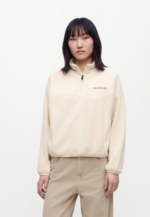 Jeune personne aux cheveux noirs raides portant un sweat crème à fermeture éclair quart et un pantalon beige, debout devant un fond blanc uni.