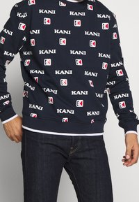 Sudadera azul marino con un patrón repetido de texto blanco "KANI" y acentos de logotipo geométrico en rojo y blanco, con puños y cuello acanalados.