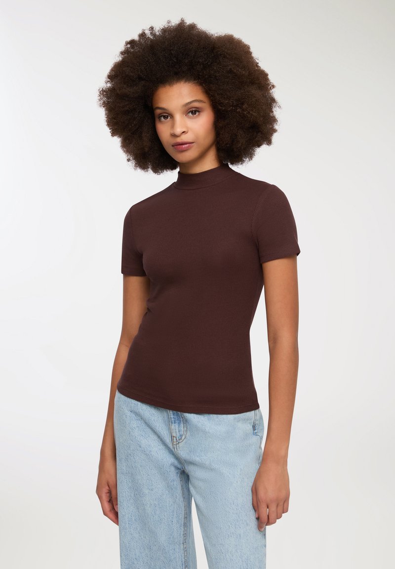 Brązowa, żeberkowana bluzka z krótkim rękawem i stójką. Tkanina jest gładka i dopasowana, w połączeniu ze jasnoniebieskimi, luźnymi dżinsami.