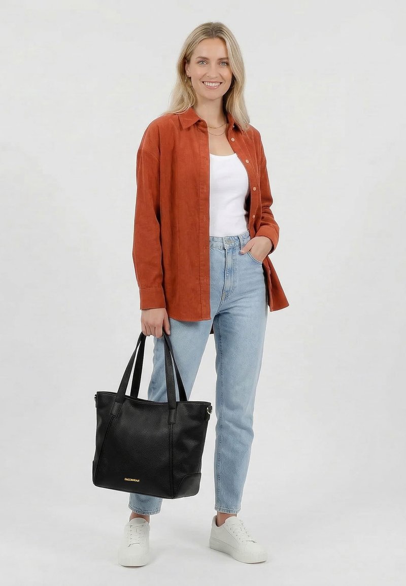 Femme souriante portant une chemise rouille, un top blanc, un jean bleu clair, des baskets blanches, tenant un grand sac fourre-tout noir avec des lettres dorées.