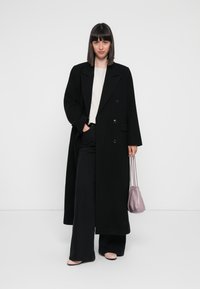 Cappotto nero doppiopetto con ampi risvolti, maniche lunghe e vestibilità rilassata, abbinato a una blusa chiara e pantaloni neri a gamba larga. Piccola borsa rosa.