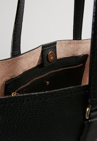 Zwarte leren tote bag met een gestructureerd uiterlijk, zachte beige suede binnenkant, voorzien van een binnenzak met ritssluiting en goudkleurige rits en hardware.