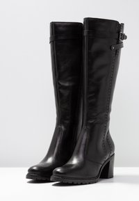 Botas de piel negra hasta la rodilla con una textura suave, con un detalle de hebilla y un tacón grueso, exhibidas sobre un fondo liso.