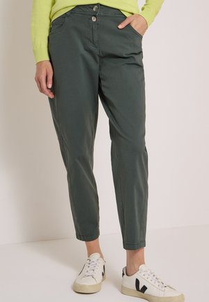 Personne portant un pantalon décontracté vert foncé et des baskets blanches avec un logo V noir, debout avec une main dans la poche sur un fond uni.