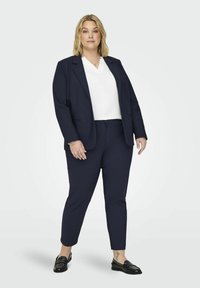 Tums zila žakete un atbilstošas bikses ar baltu krekli. Žakete ir taisna piegriezuma, bet bikses ar faktūru. Melni apavi ar sudraba akcentiem.