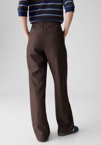 Bruine broek met wijde pijpen en een gladde textuur, voorzien van een elastische tailleband en achterzakken. Gecombineerd met een blauwe trui en sandalen.