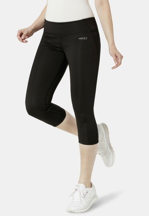 Frau in schwarzen Capri-Leggings und weißen Turnschuhen, die mit einem leicht angewinkelten Bein steht.