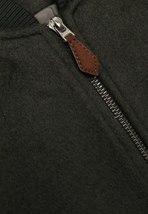 Tamnozeleno zip-up jakna s srebrnim zatvaračem i smeškim kožnim povlačnim tabom. Tkanina deluje teksturirano i meko.