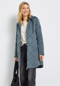 Vadderad lång kappa i dämpad blå färg, med huva, framdragkedja och dragsko. Tyget har en slät yta med diamantmönster.