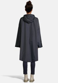 Personne avec les cheveux relevés en chignon, portant un long imperméable sombre avec une capuche, des leggings noirs et des bottines claires, se tenant de dos.