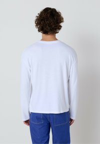 Bijela košulja dugih rukava opuštenog kroja, s okruglim izrezom i mekanom teksturom. Uparena s plavim hlačama koje prikazuju detalje šivanja.