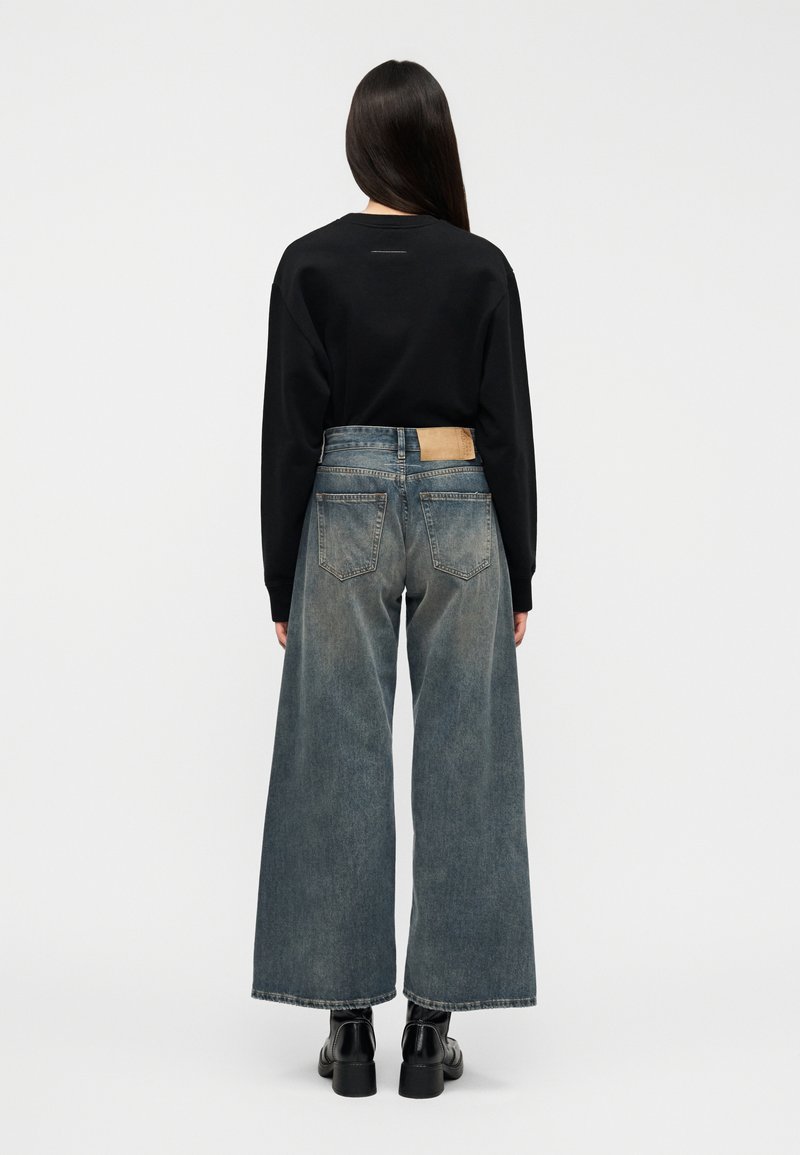 Haut noir à manches longues associé à un jean en denim bleu à jambes larges. Le jean présente une teinte claire et un écusson en cuir camel distinctif à l'arrière.