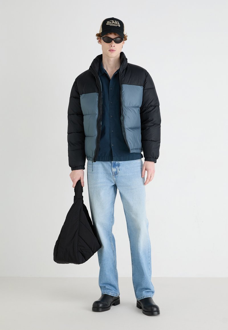 Doudoune avec des blocs de couleur noir et gris, chemise sombre, jean bleu clair, sac noir et bottes noires massives. Texture mate et design ajusté.
