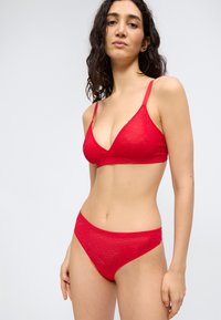 Γυναίκα με μακριά σκούρα μαλλιά που φοράει σετ κόκκινα δαντελένια σουτιέν και εσώρουχα, στέκεται μπροστά σε απλό φωτεινό φόντο.