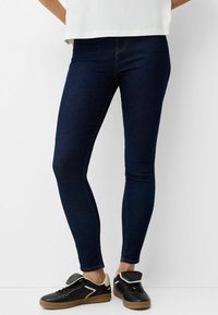 Tunna mörkblå jeans i denim, med en slät textur och traditionell femficksdesign, tillsammans med svarta sneakers.