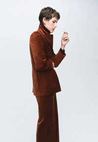 Giacca in velluto marrone con colletto alto, abbinata a pantaloni coordinati. Il tessuto presenta una texture morbida e una vestibilità sartoriale. Silhouette pulita e moderna.