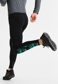 Leggings de sport noirs avec un motif géométrique turquoise sur le bas de la jambe. Le tissu est lisse et ajusté, accompagné de chaussures de sport noires.