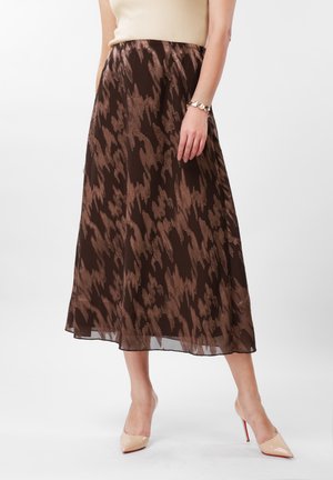 Smeđa midi suknja s uzorkom nošena s bež topom bez rukava i golim visokim potpeticama na ženi koja stoji na jednobojnoj pozadini.