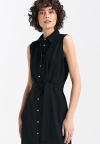 Czarna sukienka bez rękawów z kołnierzykiem, z zapięciem na guziki z przodu i związanym paskiem z materiału; wykonana z gładkiego, lekkiego materiału.
