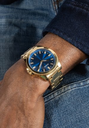 Primo piano di un polso con un orologio in metallo dorato con quadrante blu e indicatori delle ore dorati, abbinato a una manica blu scuro e jeans di denim.